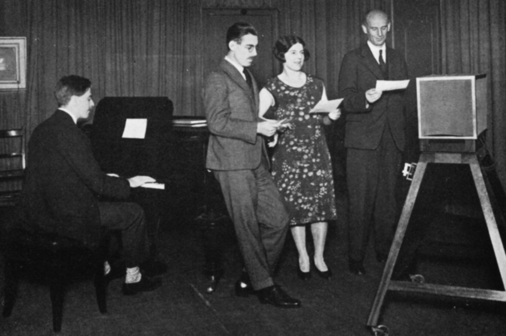 Three people read into a microphone; a man sits at a piano