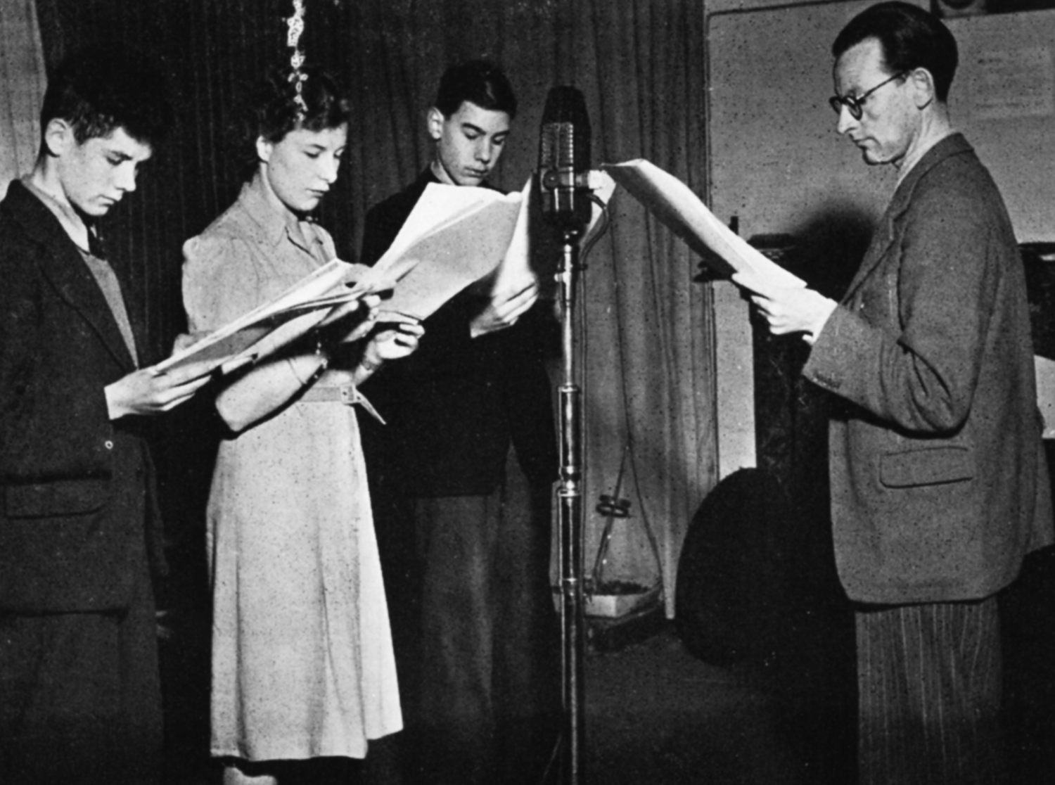 Four people hold scripts around a microphone