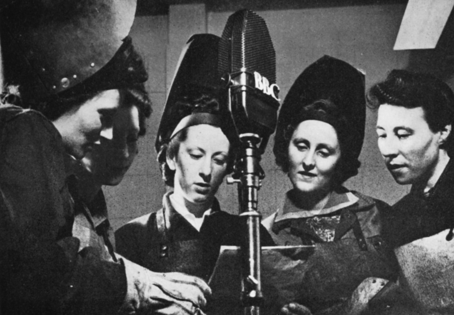 Five women around a microphone, three with welding visors